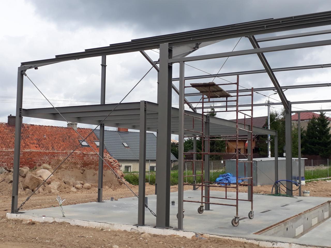 Szkielet hali stalowej w trakcie budowy, widoczny fundament, rusztowanie i fragment zniszczonego budynku w tle.