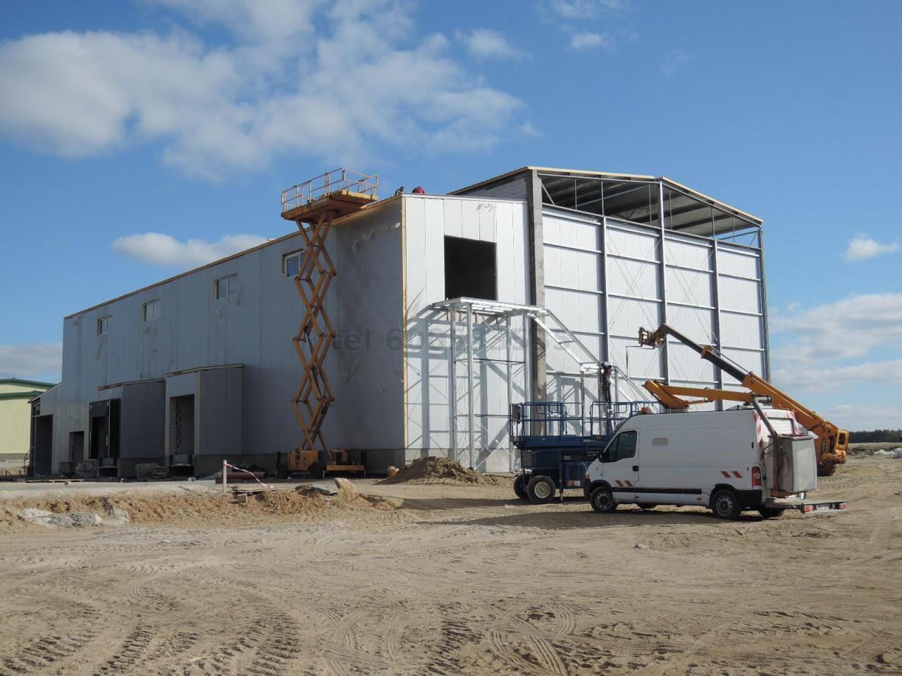 Wznoszenie hali z konstrukcji stalowej, widoczne rusztowania i podnośniki, częściowo otynkowana ściana boczna z bramami wjazdowymi, plac budowy z piaskiem.