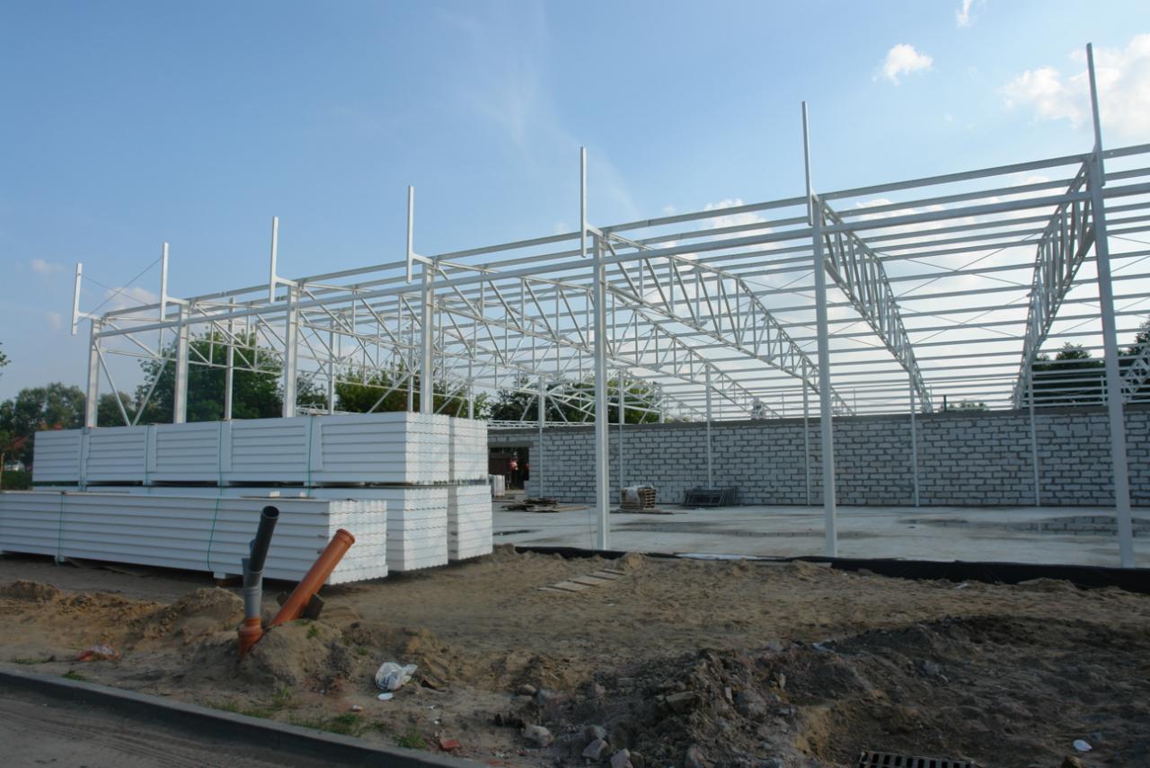 Wznoszenie szkieletu hali z białych profili stalowych, widoczne przygotowane płyty warstwowe i fragment murowanej ściany.