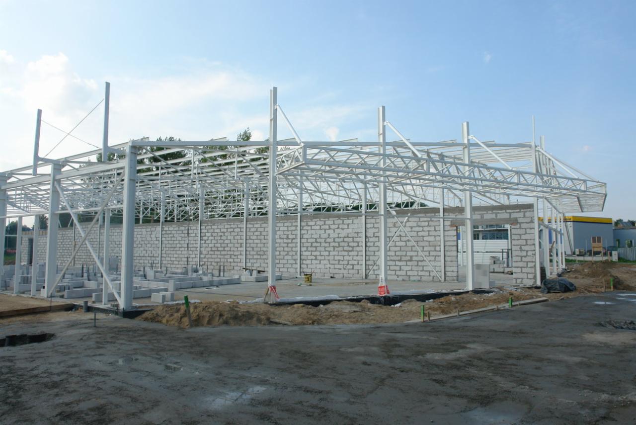 Konstrukcja stalowa hali w trakcie budowy, widoczne białe słupy i kratownica dachu, częściowo wymurowane ściany z bloczków, przygotowany teren pod wylewkę, w tle budynek w kolorze żółto-szarym.