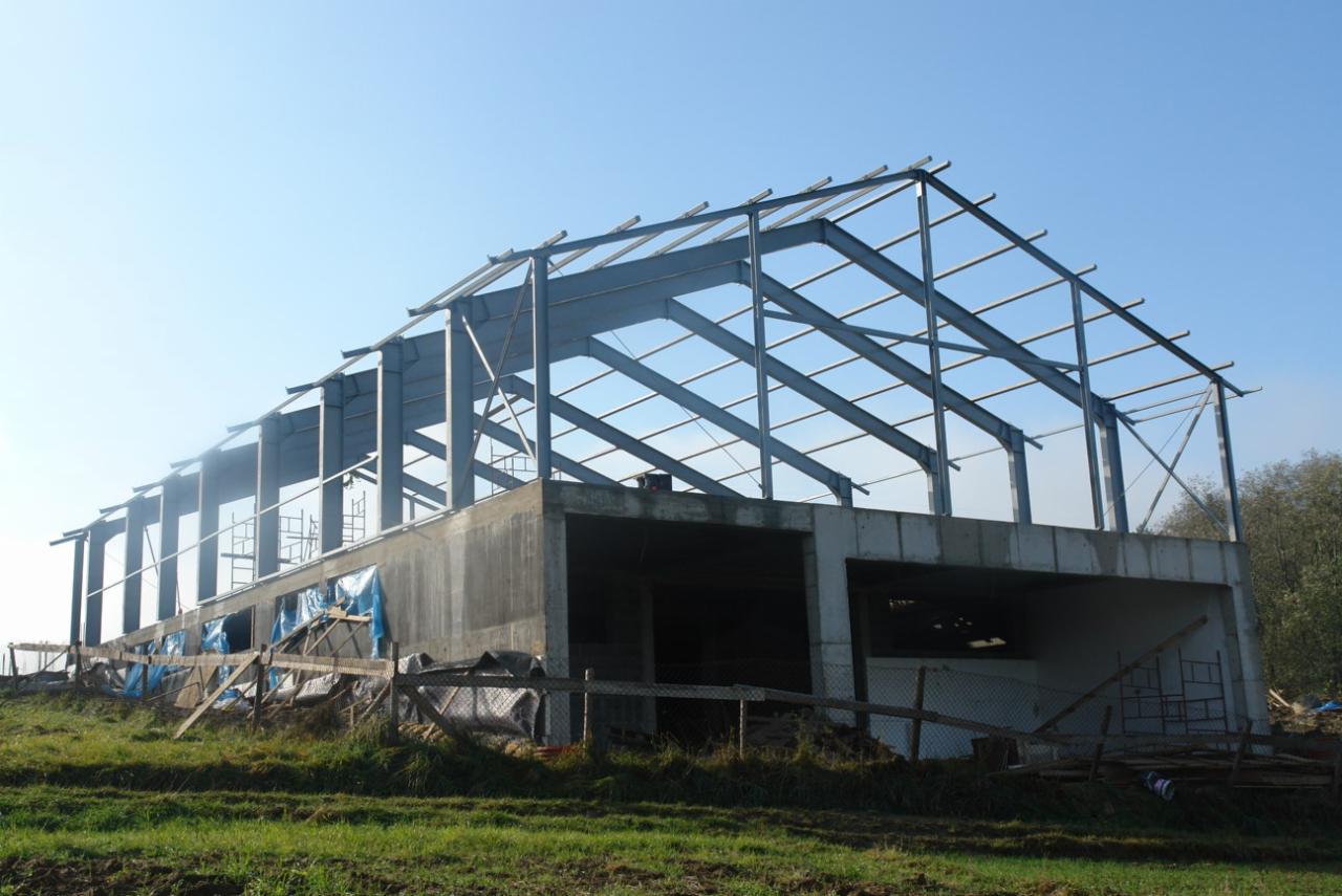 Wznoszona hala z betonowym fundamentem i stalową konstrukcją szkieletową dachu, widok z zewnątrz na tle błękitnego nieba.