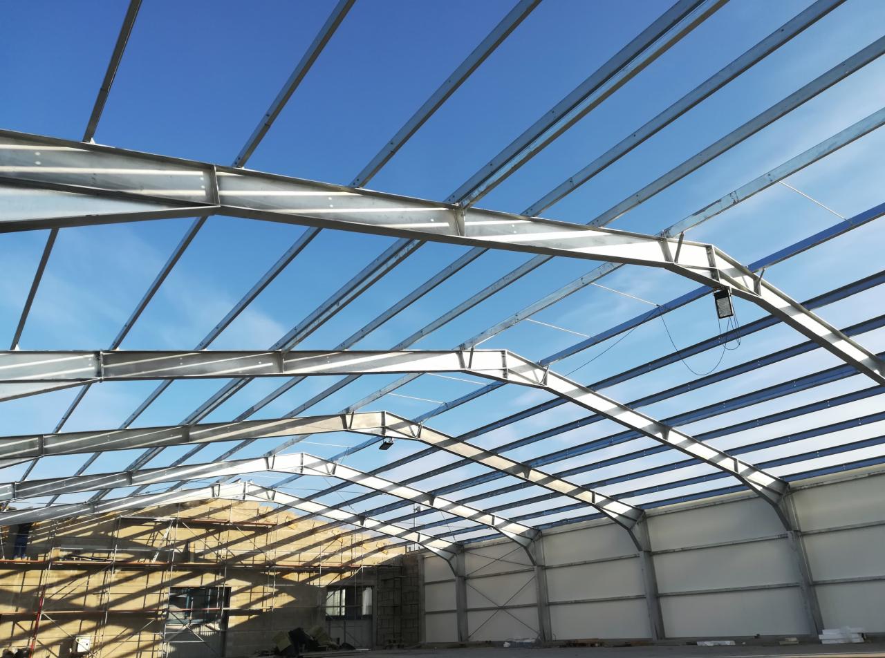 Wnętrze hali w trakcie budowy, widoczne stalowe elementy konstrukcyjne pod jasnym, błękitnym niebem, ściany boczne częściowo obudowane płytami warstwowymi, a w tle rusztowania przy ścianie szczytowej.