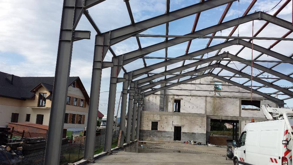 Wznoszenie konstrukcji stalowej hali na tle budynku mieszkalnego, widoczne słupy i elementy dachu, biały samochód dostawczy w rogu kadru.