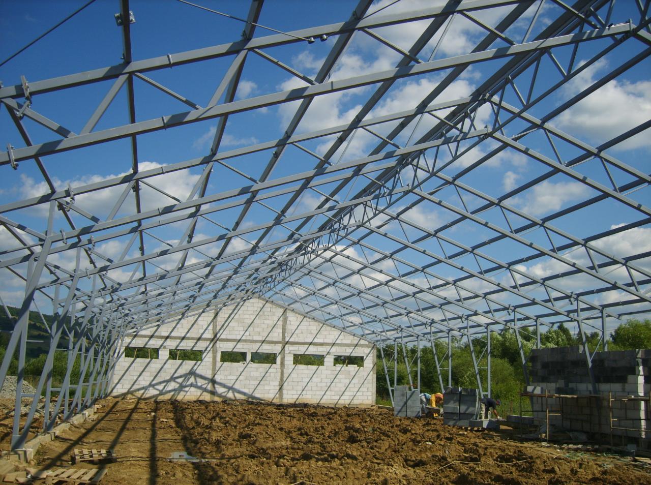 Wnętrze budowanej hali z widoczną stalową konstrukcją dachu na tle błękitnego nieba, częściowo wzniesione ściany z bloczków betonowych, oraz dwóch pracowników w oddali.