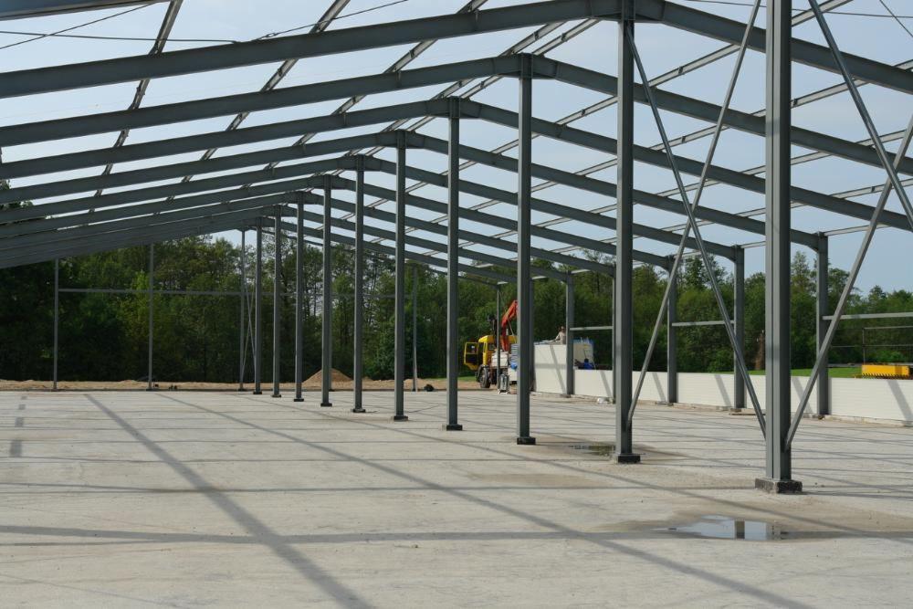 Wnętrze hali w trakcie budowy, widoczne stalowe słupy i konstrukcja dachu, betonowa posadzka, w tle las i częściowo zamontowane panele ścienne oraz żółty dźwig.