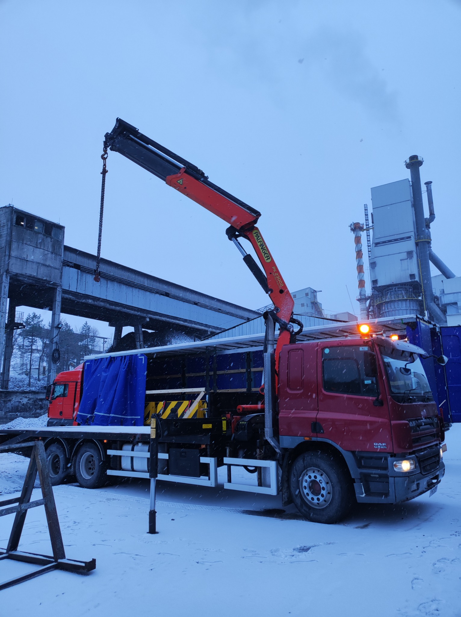 Czerwona ciężarówka z pomarańczowym dźwigiem Palfinger PK 23002 podczas zaśnieżonej pogody na tle przemysłowego budynku.