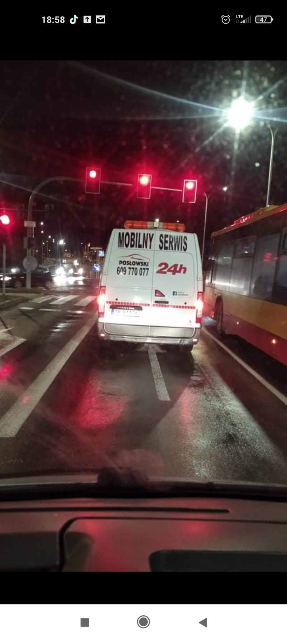Tył białego samochodu dostawczego z napisem 'Mobilny Serwis 24h' stojącego na mokrej jezdni przed światłami w nocy, widziany z wnętrza innego pojazdu.