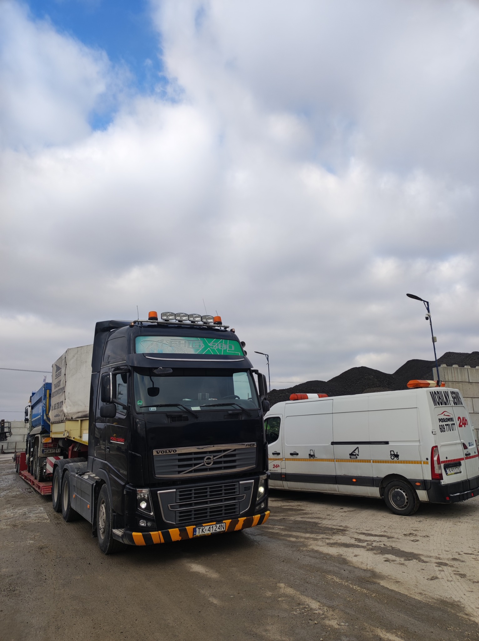 Czarny ciągnik siodłowy Volvo z naczepą przewożącą ładunek pod plandeką, zaparkowany obok białego busa serwisowego z napisem 'Mobilny Serwis 24h', na placu z hałdą węgla w tle.