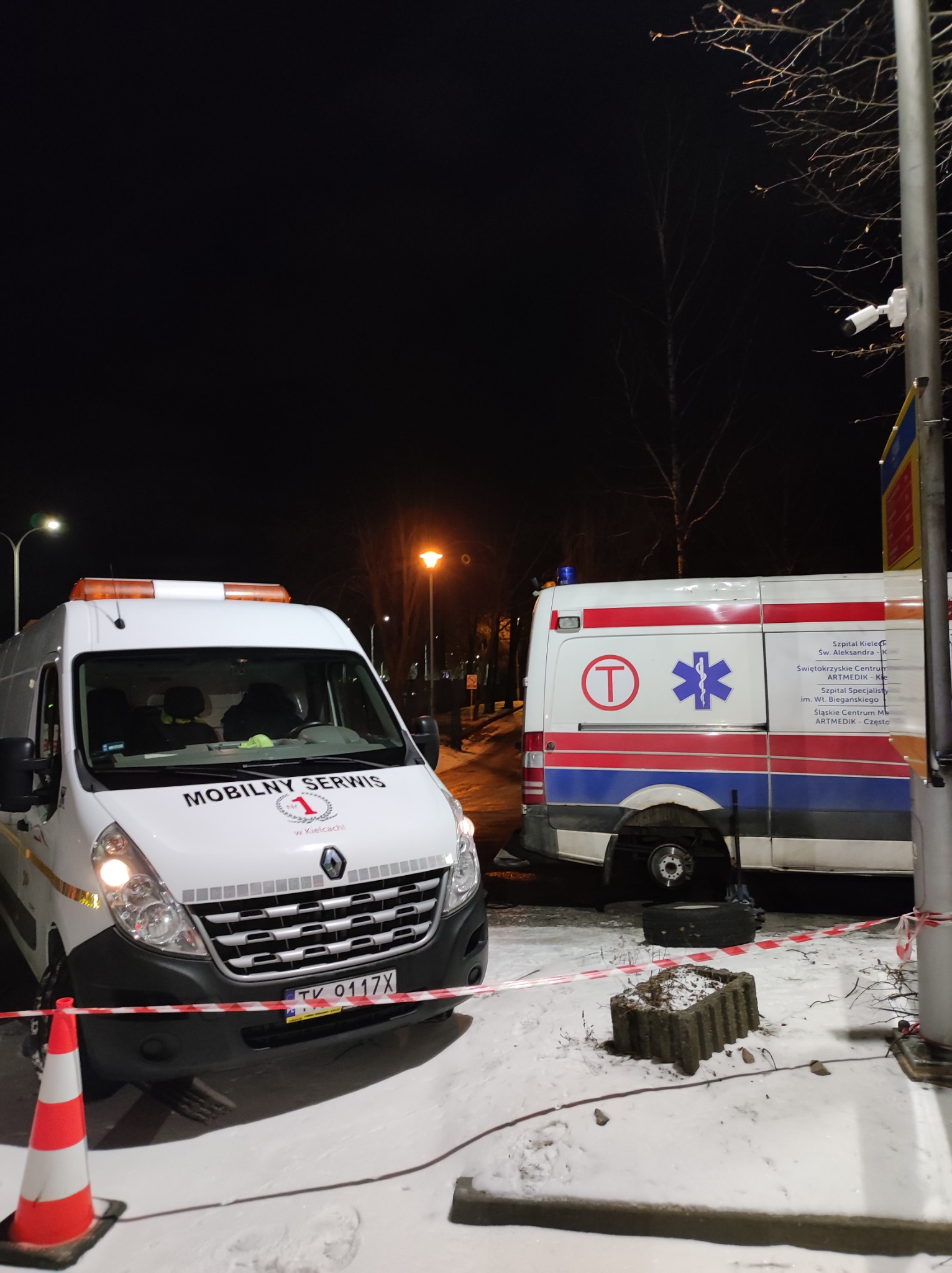 Dwa pojazdy: biały van z napisem Mobilny Serwis z przodu i ambulans w tle, oba stoją na śniegu w nocy, oddzielone taśmą ostrzegawczą, z widocznym brakiem opony w ambulansie.