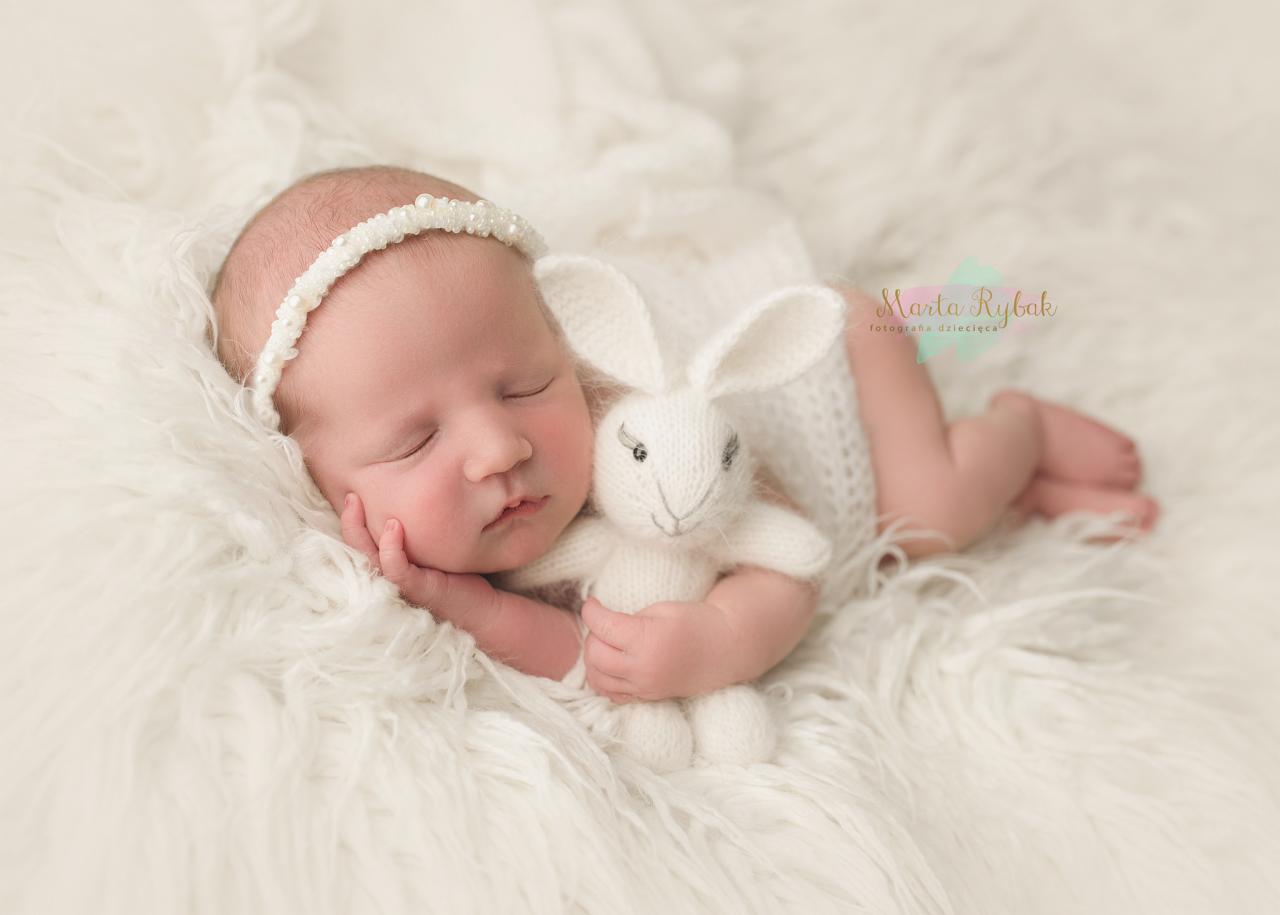 Śpiący noworodek z białą opaską na głowie, leżący na puszystym, białym tle, trzymający w rączkach pluszowego, białego króliczka.