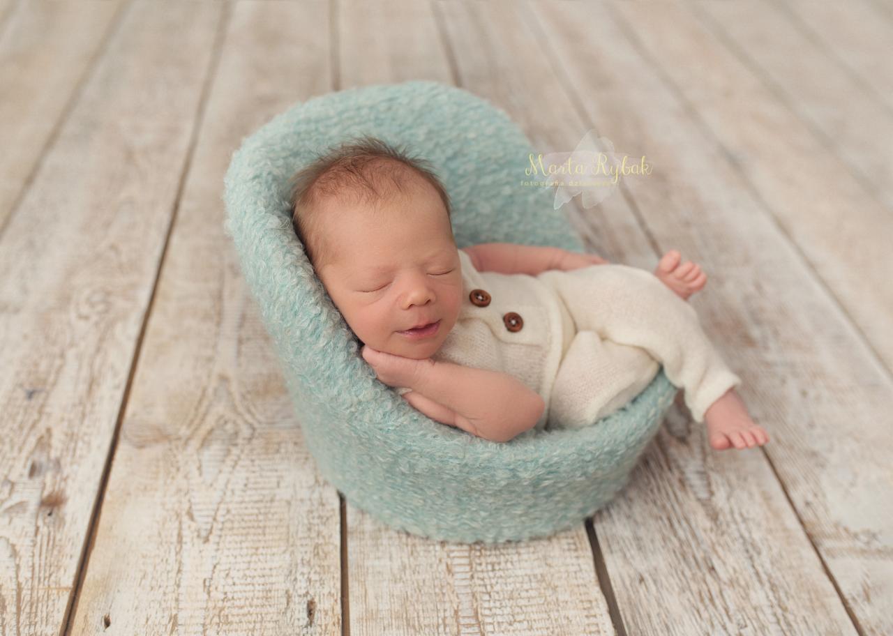 Śpiący noworodek w kremowym ubranku z drewnianymi guzikami, ułożony w małym, turkusowym fotelu z dzianiny na tle z desek.