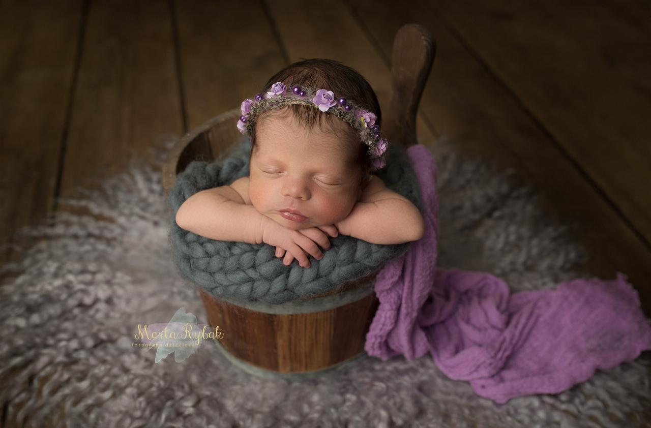 Śpiący noworodek w drewnianym wiaderku, otulony szarą wełną i fioletowym materiałem, z delikatnym wiankiem z fioletowych kwiatów na głowie, na tle drewnianej ściany i futrzanej podkładki.