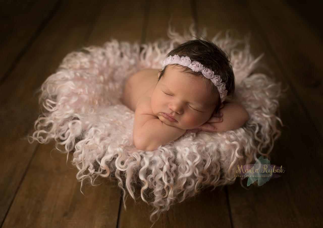 Śpiąca noworodka z ciemnymi włosami i różową opaską z kwiatami na głowie, ułożona na puszystym, różowym kocyku z kręconymi włóknami na drewnianym tle.