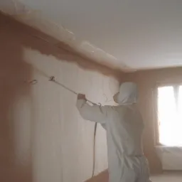 Pracownik w kombinezonie ochronnym natryskowo nakłada gładź na ścianę w pokoju, zabezpieczone folią obrzeża sufitu, widoczne okno i lampa.