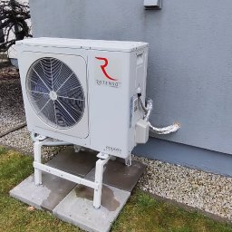 QNA Ochrona Jakub Maciej Kuna - Biała zewnętrzna jednostka pompy ciepła Rotenso Aquami, zamontowana na metalowym stelażu na betonowych płytach, przy szarej ścianie budynku z widocznymi przewodami w izolacji.