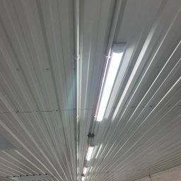 QNA Ochrona Jakub Maciej Kuna - Wnętrze hali z zamontowanym oświetleniem: rzędy lamp liniowych na metalowym, trapezowym suficie, widoczne rury instalacyjne i punkty mocowania.