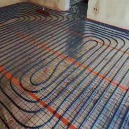 Rozłożone, niebieskie rury ogrzewania podłogowego na siatce, widoczne pomarańczowe elementy mocujące, przygotowane do zalania wylewką w surowym wnętrzu.