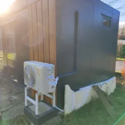 Zewnętrzna jednostka klimatyzacji marki Rotenso zamontowana na białym stelażu przy elewacji budynku w odcieniach szarości i drewna. Widoczne przewody i rynna odprowadzająca wodę. W tle trawa...