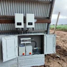 Dwa białe inwertery fotowoltaiczne zamontowane pod blachą falistą, z otwartą skrzynką elektryczną zawierającą zabezpieczenia i okablowanie, na tle błotnistego terenu.