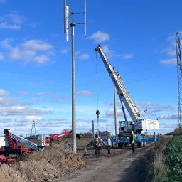 QNA Ochrona Jakub Maciej Kuna - Montaż pionowej turbiny wiatrowej na polu, z użyciem dźwigu Herkules i podnośnika Manitou, widoczni pracownicy w kamizelkach odblaskowych.