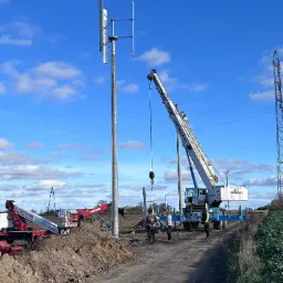 Montaż pionowej turbiny wiatrowej na polu, z użyciem dźwigu Herkules i podnośnika Manitou, widoczni pracownicy w kamizelkach odblaskowych.