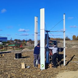 QNA Ochrona Jakub Maciej Kuna - Montaż turbiny wiatrowej pionowej osi obrotu na polu, dwóch monterów przy pracy, widoczny dźwig, błękitne niebo.