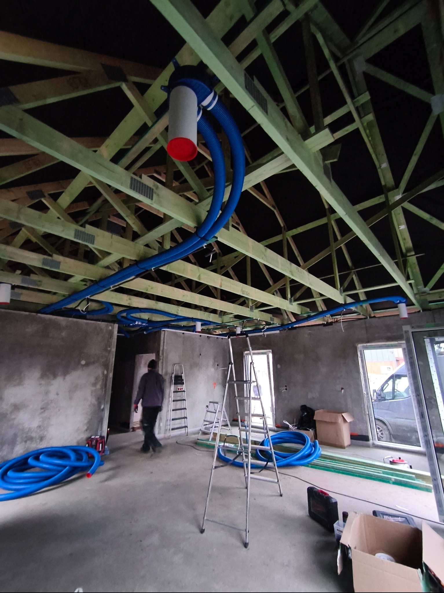 Surowy stan budynku z widoczną instalacją wentylacyjną pod sufitem, niebieskie rury rozprowadzone wzdłuż drewnianej konstrukcji dachu, drabiny i materiały budowlane na podłodze.