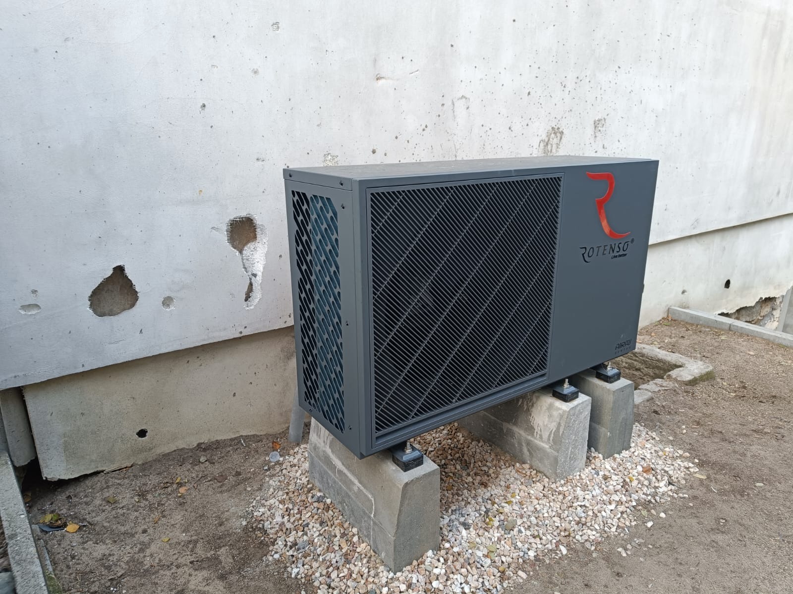 Szara pompa ciepła Rotenso ustawiona na betonowych bloczkach przed szarą, uszkodzoną ścianą budynku, otoczona drobnymi kamyczkami.