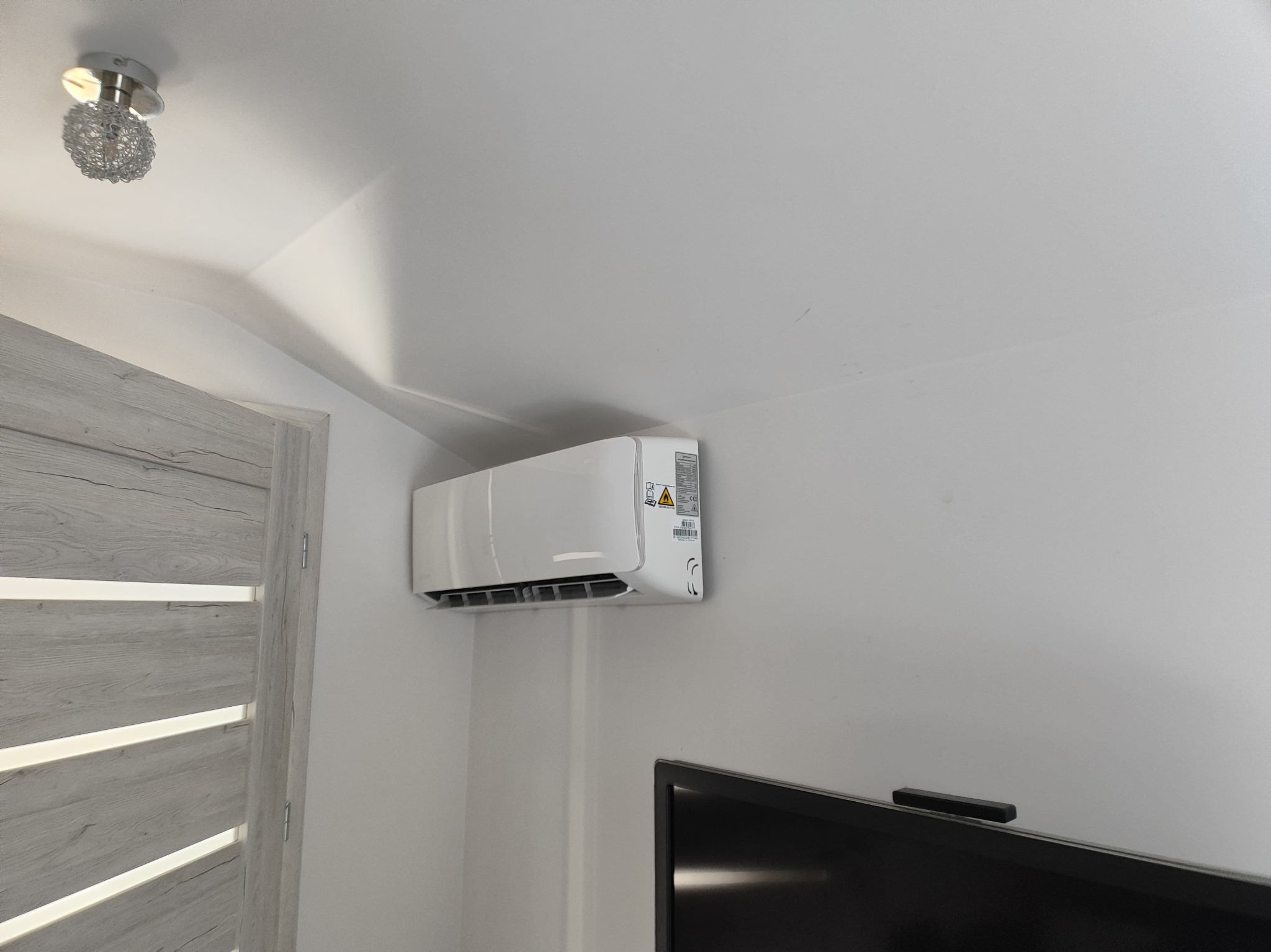 Biały klimatyzator ścienny zamontowany w rogu pokoju, widoczny fragment drzwi z dekoracyjną fakturą drewna i lampa sufitowa w stylu glamour, obok telewizor.