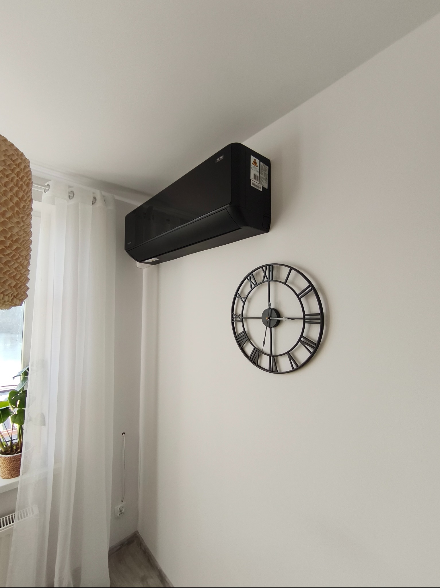 Czarny klimatyzator ścienny zamontowany w jasnym pokoju, obok dekoracyjny zegar z rzymskimi cyframi, widoczne firanki i lampa z abażurem.