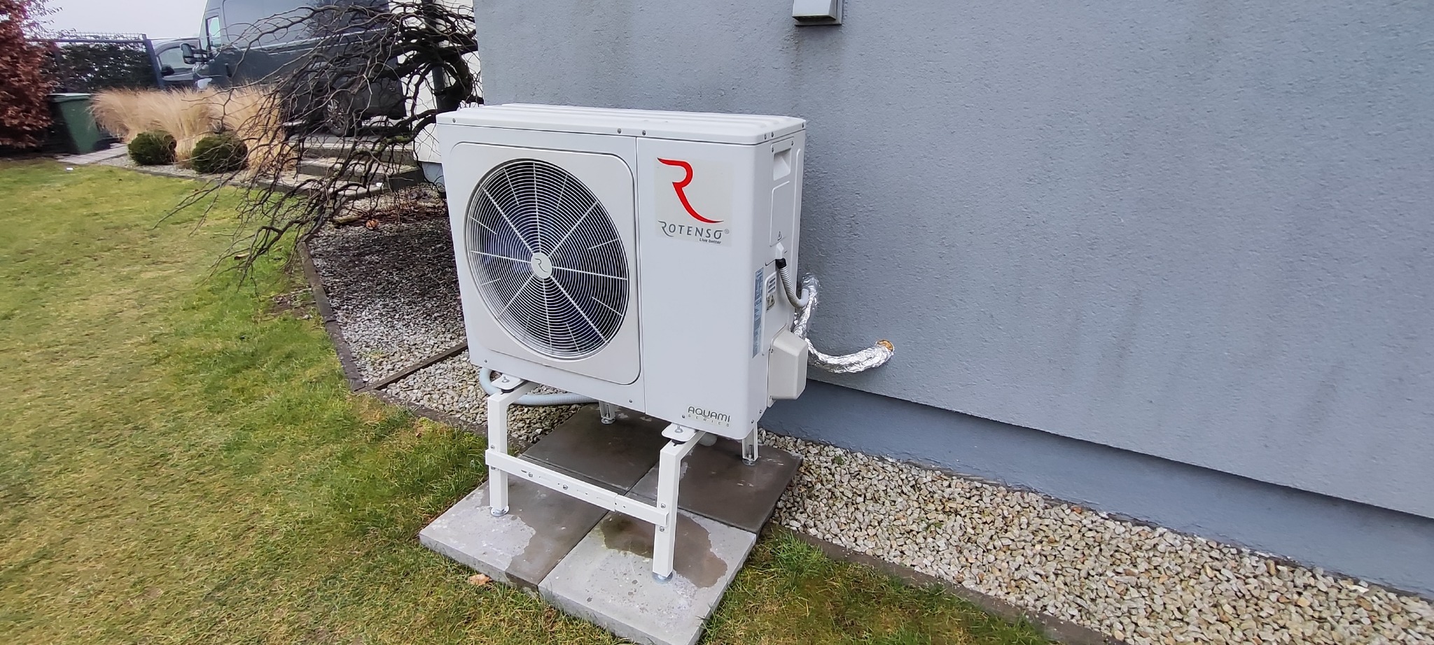 Biała zewnętrzna jednostka pompy ciepła Rotenso Aquami, zamontowana na metalowym stelażu na betonowych płytach, przy szarej ścianie budynku z widocznymi przewodami w izolacji.