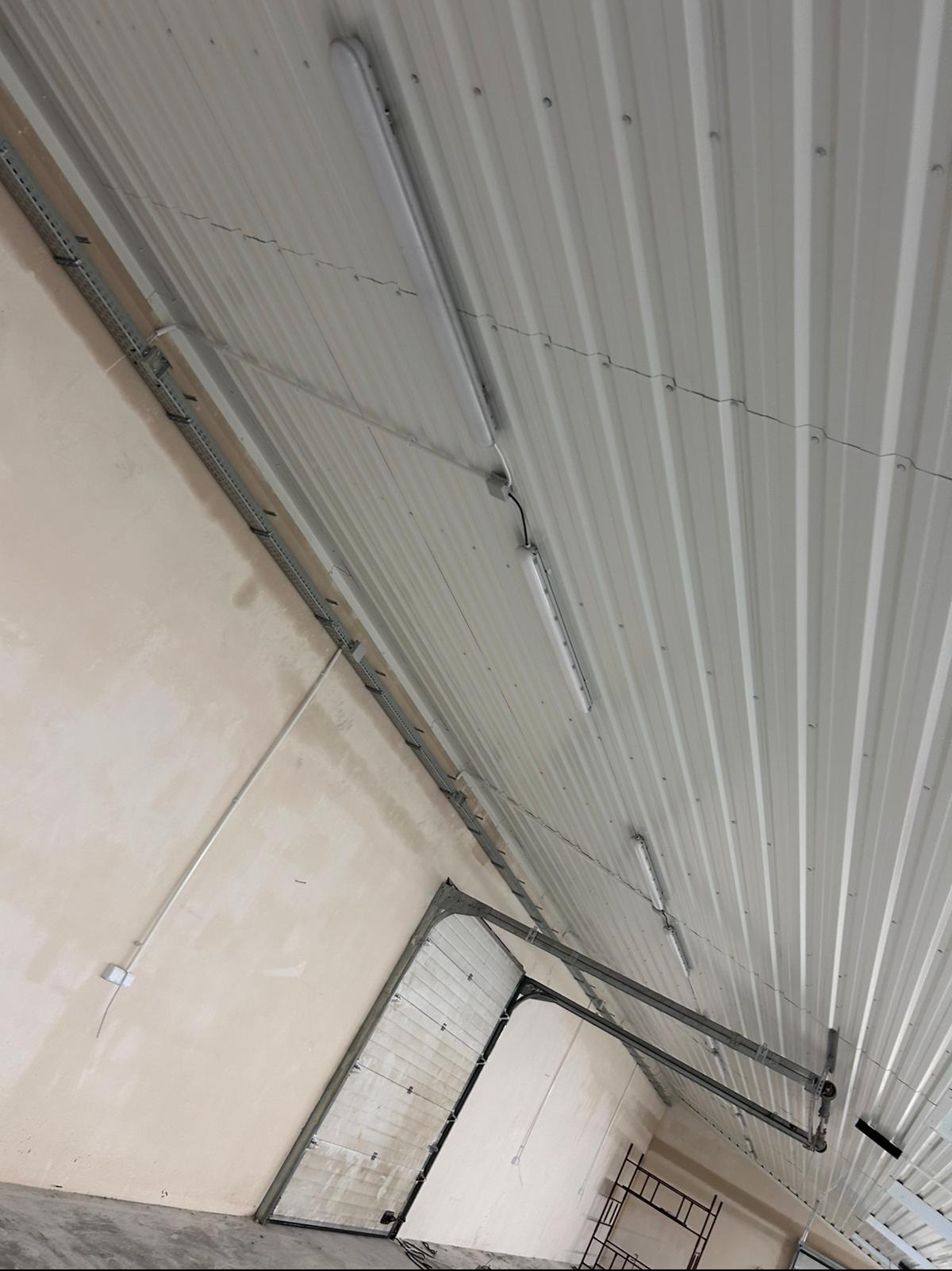 Wnętrze hali produkcyjnej z widoczną instalacją oświetleniową pod sufitem z blachy trapezowej, korytkami kablowymi przy ścianie i częściowo otwartą bramą garażową. Widoczne są również elementy...