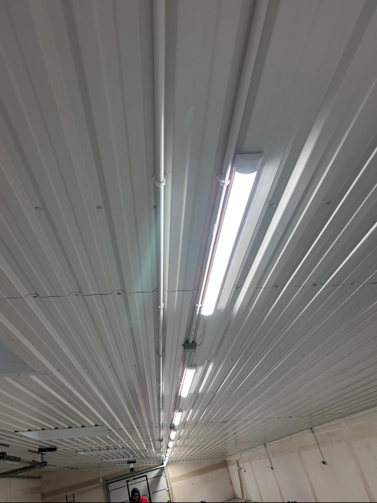 Wnętrze hali z zamontowanym oświetleniem: rzędy lamp liniowych na metalowym, trapezowym suficie, widoczne rury instalacyjne i punkty mocowania.