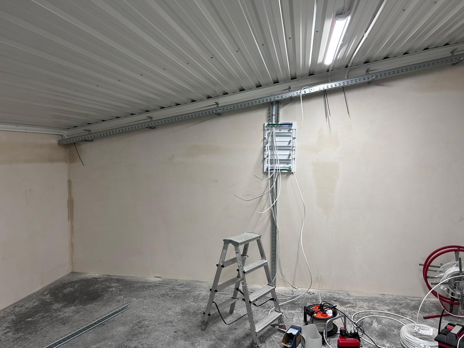 Rozdzielnica elektryczna z podłączonymi przewodami na ścianie w surowym pomieszczeniu z blachodachówką, widoczna drabina aluminiowa i zwój przewodów na podłodze.