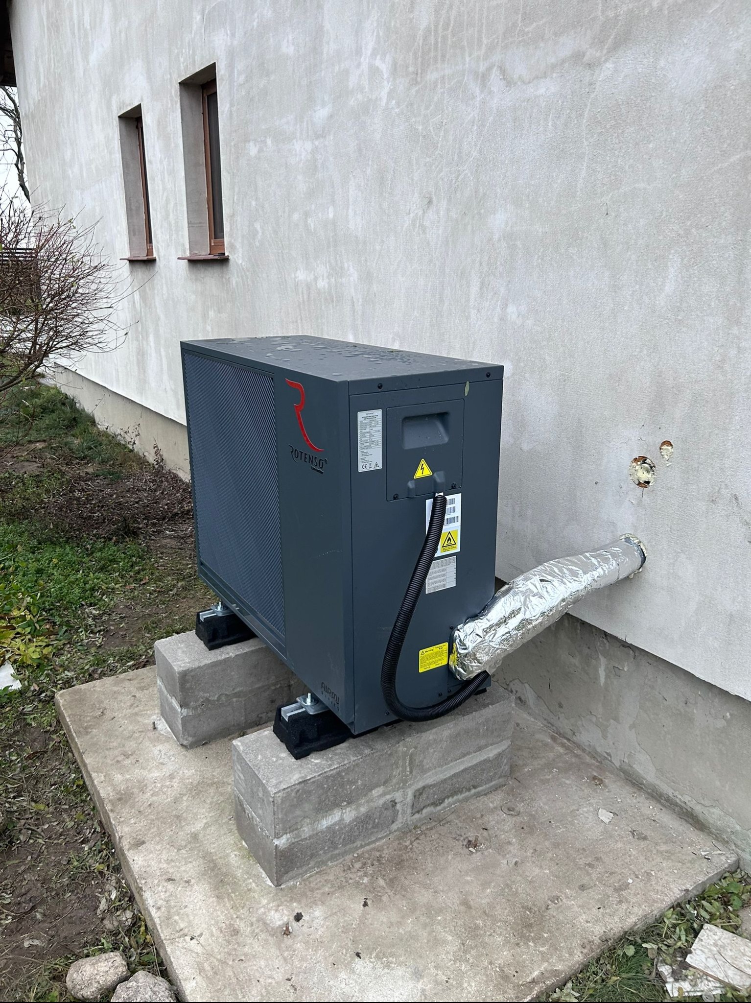 Szara pompa ciepła Rotenso umieszczona na betonowych blokach przy ścianie budynku z widocznym przewodem w izolacji aluminiowej wchodzącym do otworu w ścianie.