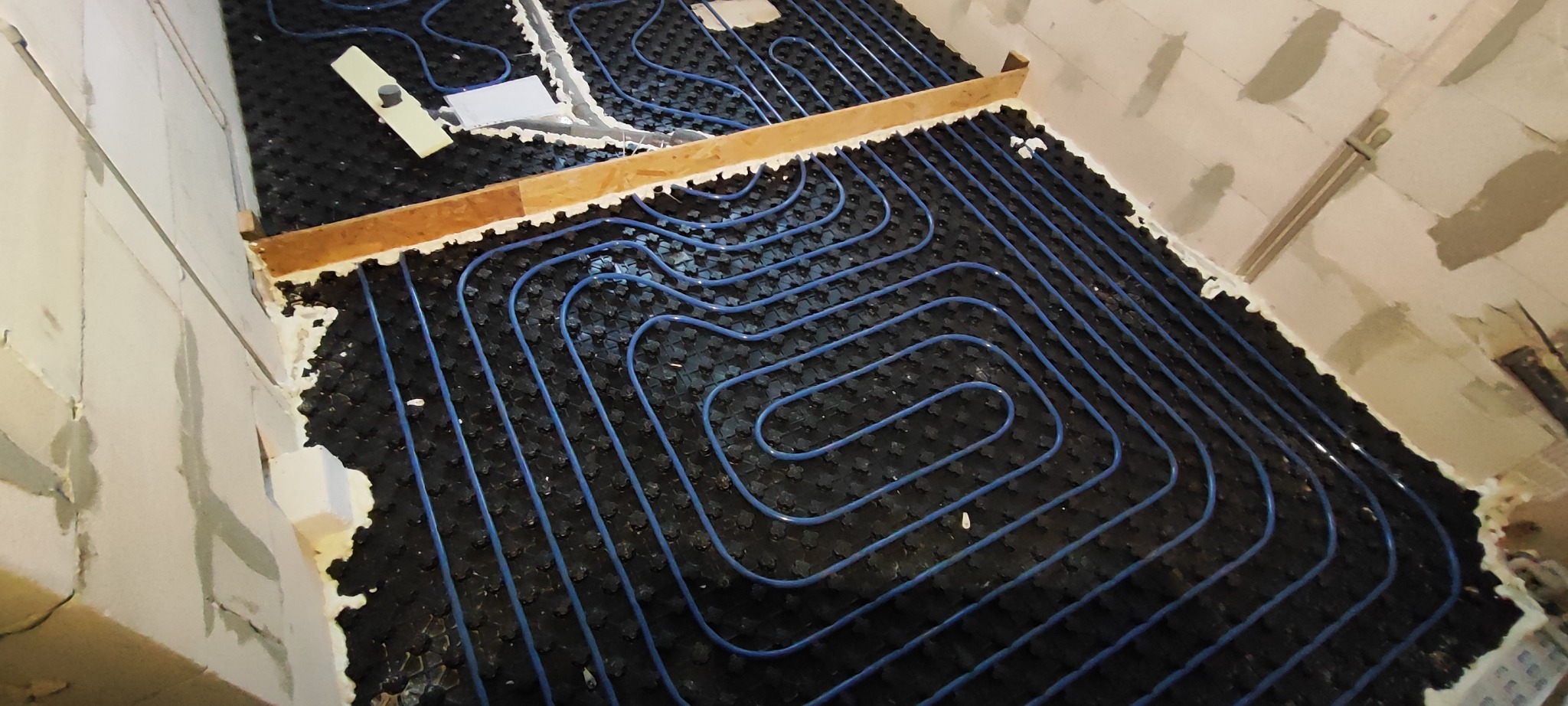 Niedokończona instalacja ogrzewania podłogowego z niebieskimi rurkami na czarnej macie izolacyjnej, widoczne fragmenty ścian w surowym stanie i drewniana listwa mocująca.