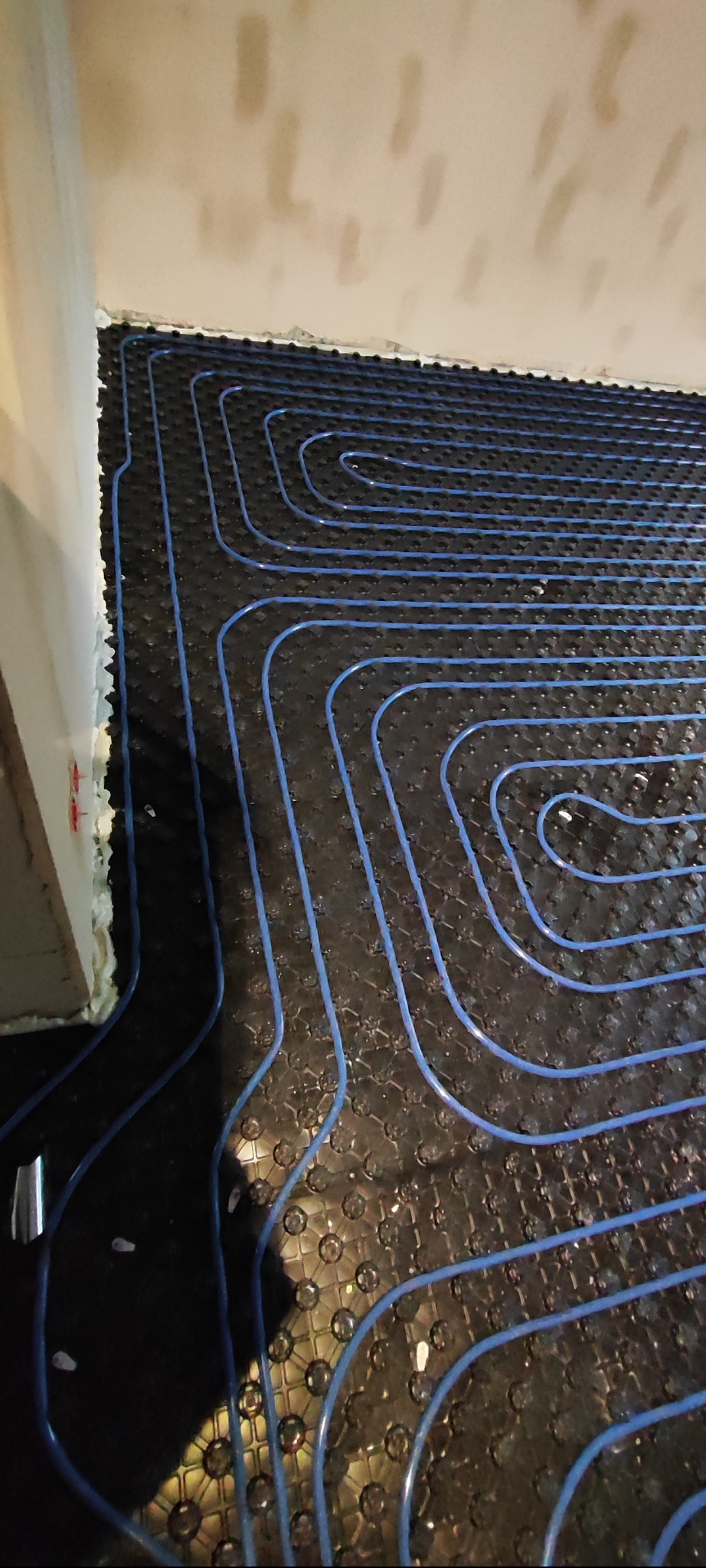 Niebieskie rurki ogrzewania podłogowego ułożone na czarnej macie izolacyjnej, widoczne fragmenty ściany i pianki montażowej.