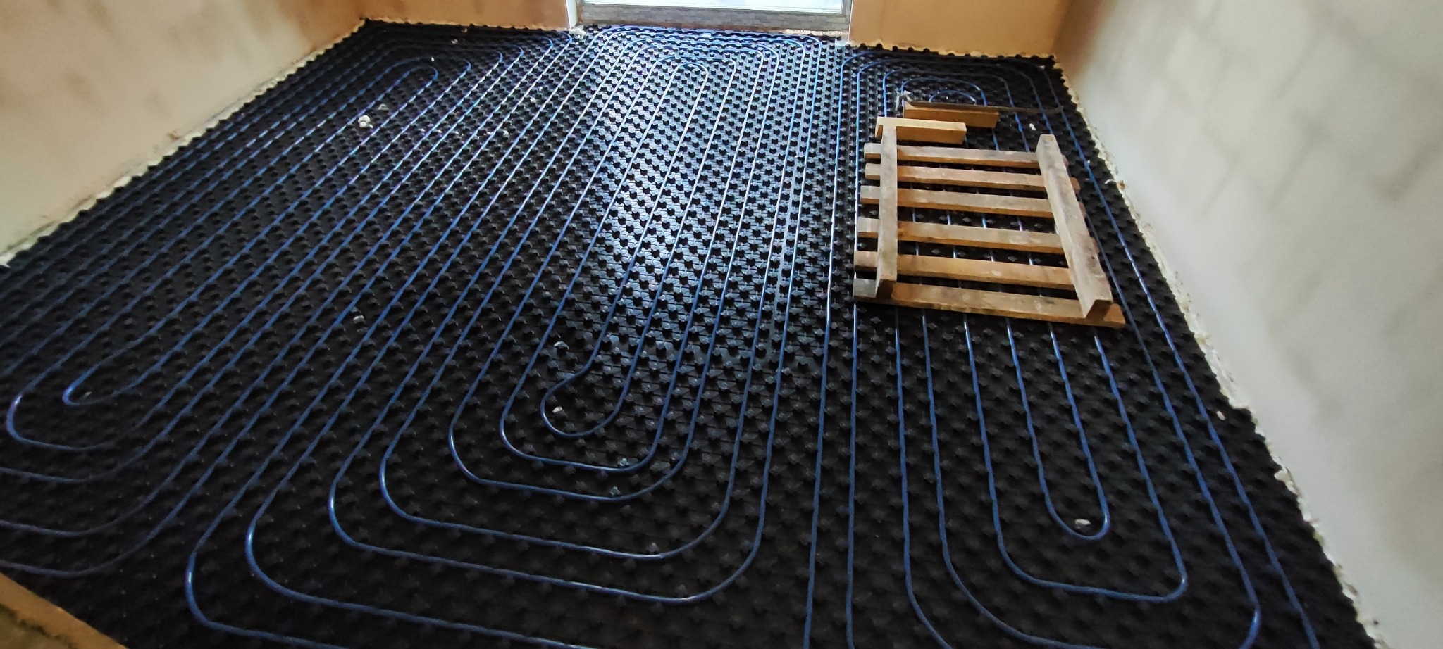 Niebieskie rury ogrzewania podłogowego ułożone na czarnej macie izolacyjnej w surowym wnętrzu, z drewnianą konstrukcją na wierzchu.