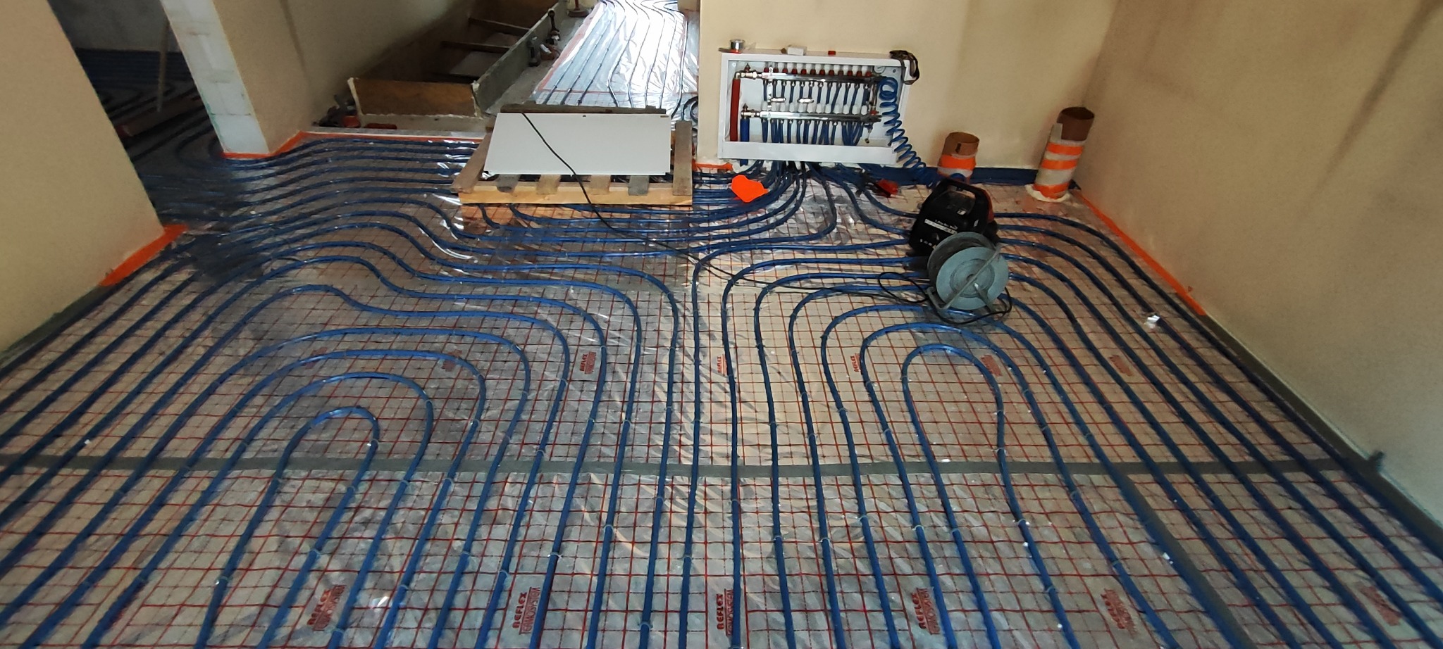 Rozłożone niebieskie rury ogrzewania podłogowego na folii z czerwoną siatką, widoczny rozdzielacz i narzędzia montażowe.