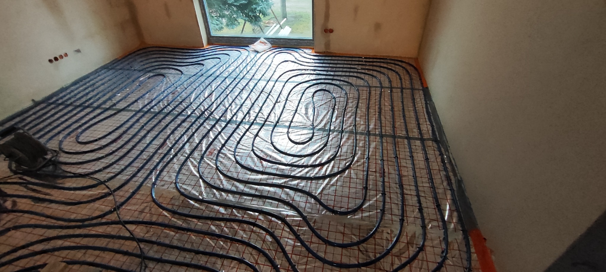 Czarny system rur ogrzewania podłogowego ułożony na siatce, pokryty folią, widok na okno i surowe ściany