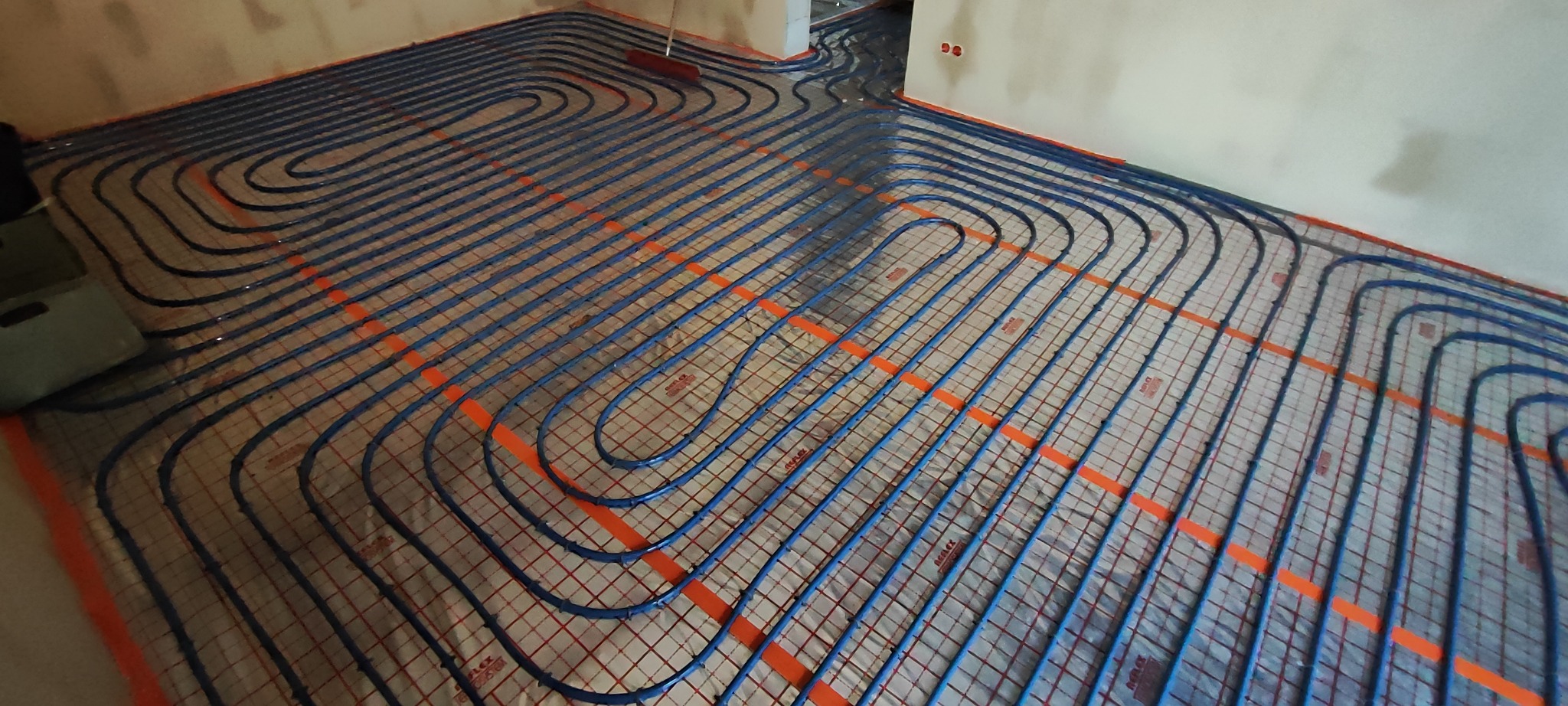 Rozłożone, niebieskie rury ogrzewania podłogowego na siatce, widoczne pomarańczowe elementy mocujące, przygotowane do zalania wylewką w surowym wnętrzu.