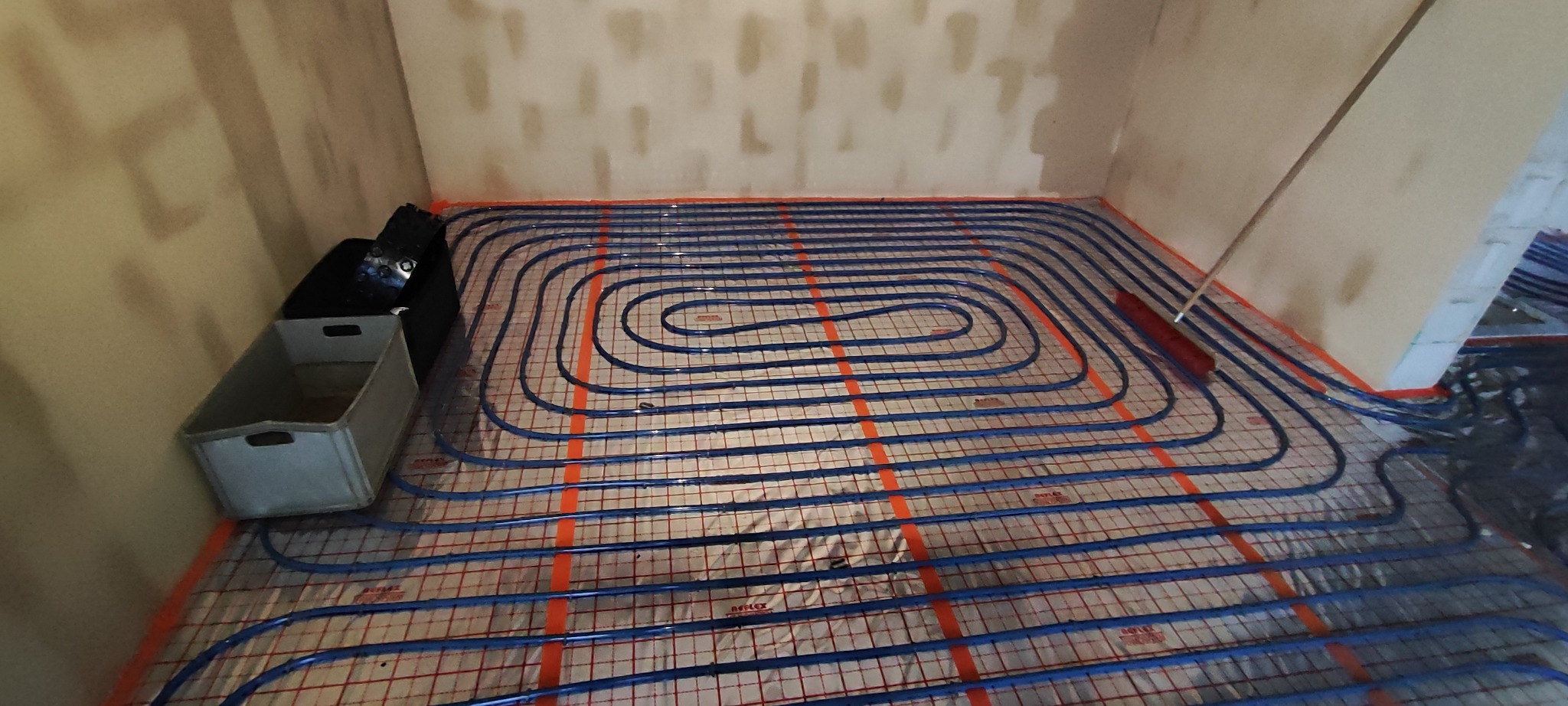 Niebieskie rury ogrzewania podłogowego ułożone spiralnie na czerwonej macie izolacyjnej w pomieszczeniu w Unisławiu, widoczna skrzynka narzędziowa i przygotowane ściany.