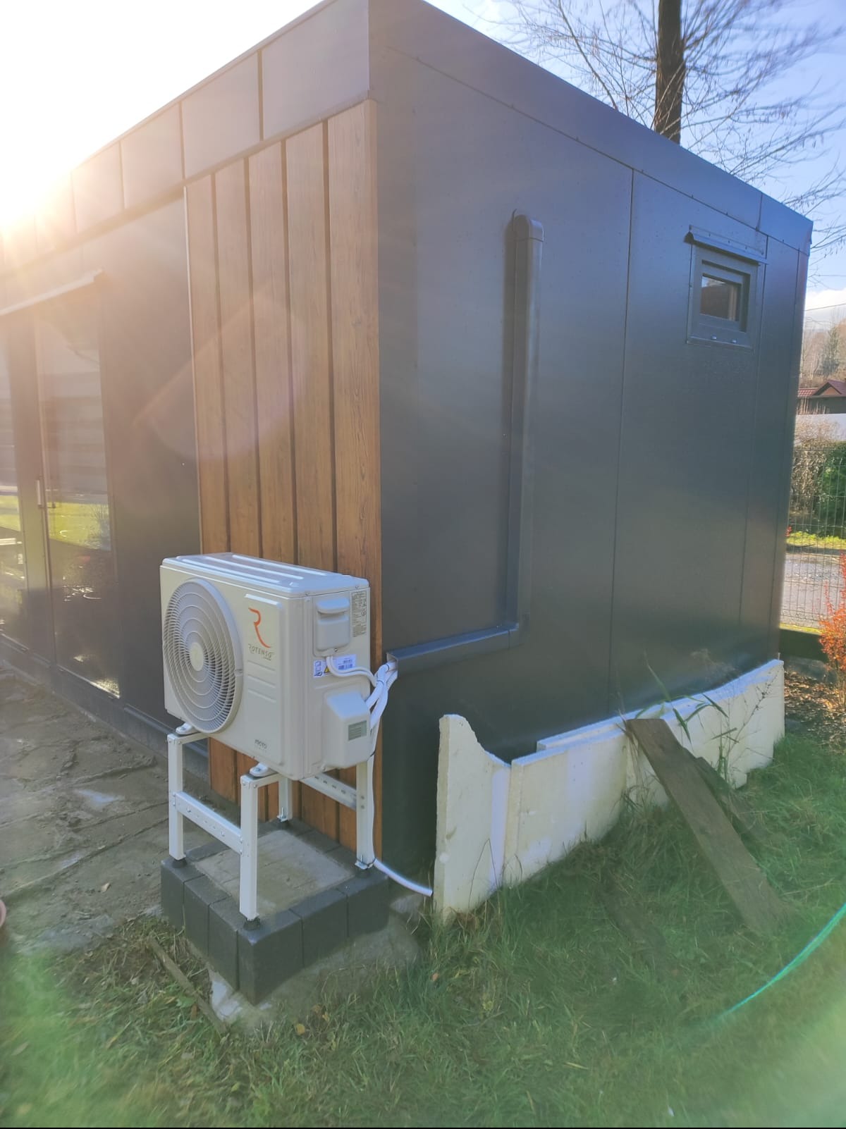 Zewnętrzna jednostka klimatyzacji marki Rotenso zamontowana na białym stelażu przy elewacji budynku w odcieniach szarości i drewna. Widoczne przewody i rynna odprowadzająca wodę. W tle trawa...