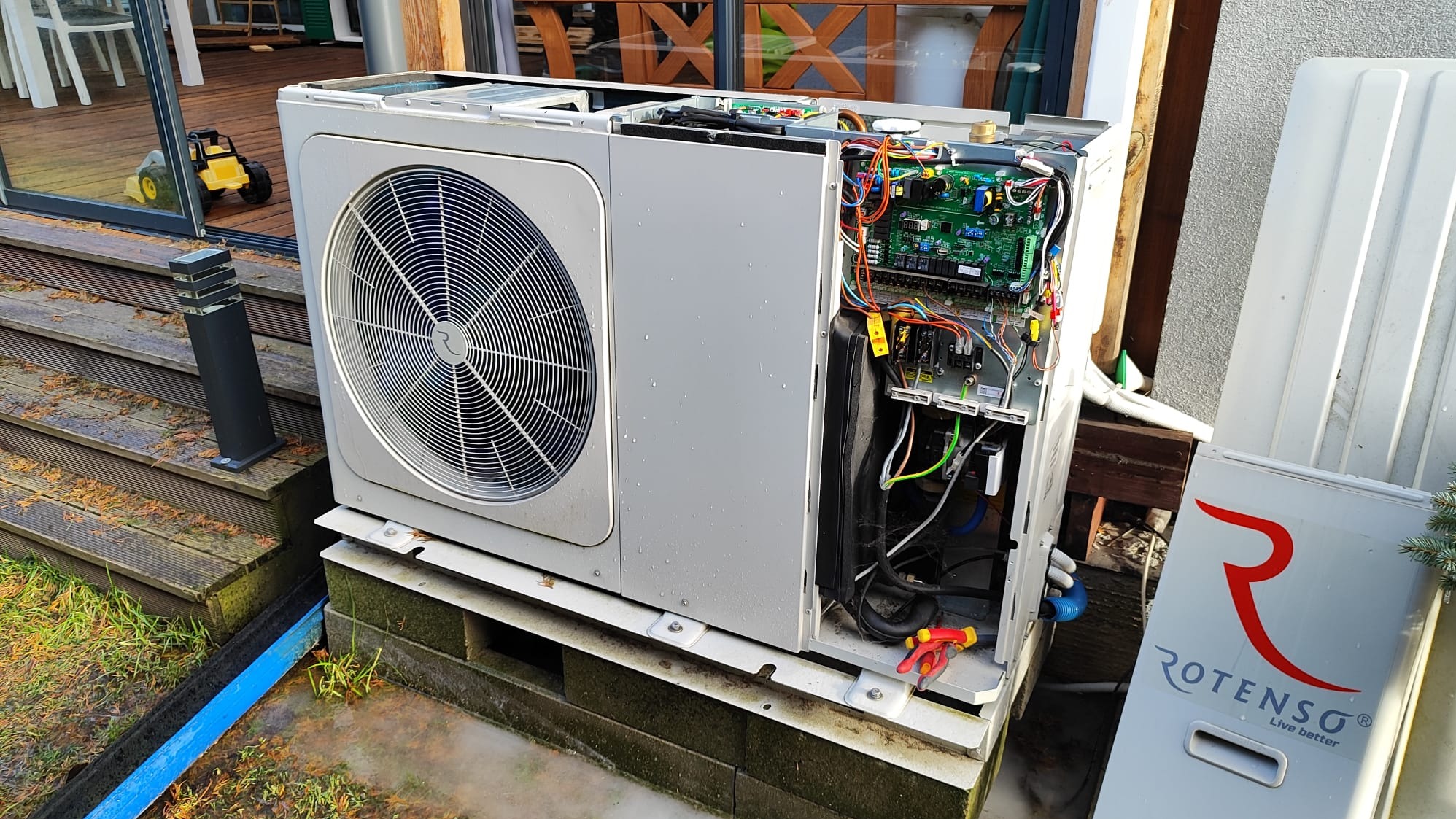 Otwarta obudowa pompy ciepła marki Rotenso na zewnątrz budynku, widoczne wewnętrzne komponenty, w tym płytka drukowana i okablowanie, obok leżą czerwono-żółte szczypce.