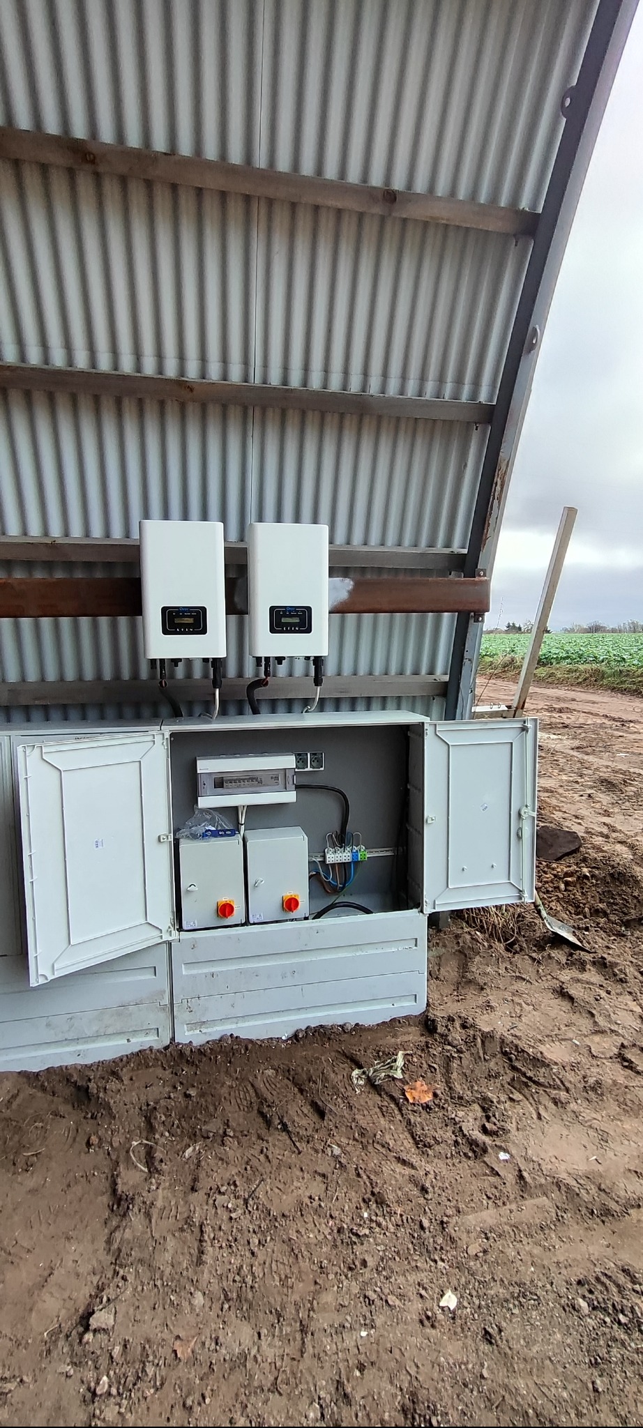 Dwa białe inwertery fotowoltaiczne zamontowane pod blachą falistą, z otwartą skrzynką elektryczną zawierającą zabezpieczenia i okablowanie, na tle błotnistego terenu.