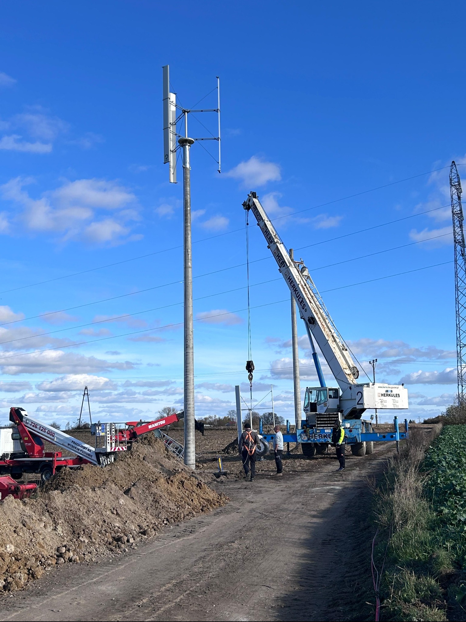 Montaż pionowej turbiny wiatrowej na polu, z użyciem dźwigu Herkules i podnośnika Manitou, widoczni pracownicy w kamizelkach odblaskowych.
