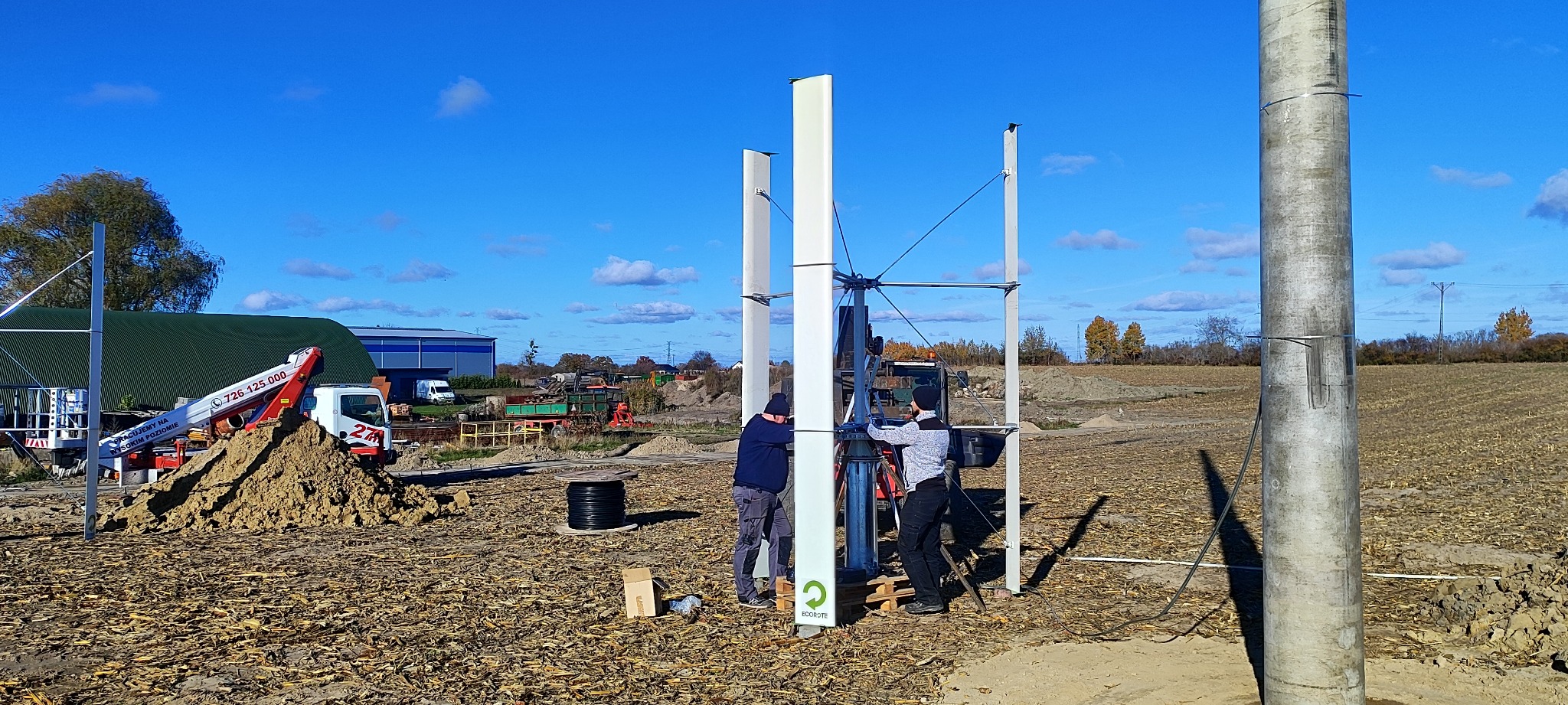 Montaż turbiny wiatrowej pionowej osi obrotu na polu, dwóch monterów przy pracy, widoczny dźwig, błękitne niebo.