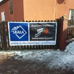 Baner reklamowy firmy 'Skała' oferującej sprzedaż i wykonawstwo ocieplania budynków energooszczędnych, zawieszony na drewnianym płocie przed budynkiem w zimowej scenerii.