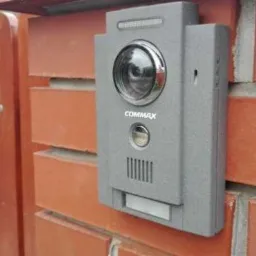 Szary panel wideodomofonu Commax zamontowany na ceglanym murze obok drewnianego ogrodzenia.