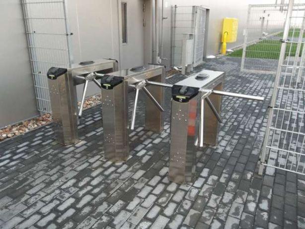 Trzy stalowe bramki obrotowe na zewnątrz budynku, ustawione na szarej kostce brukowej, z widocznymi elementami ogrodzenia w tle.
